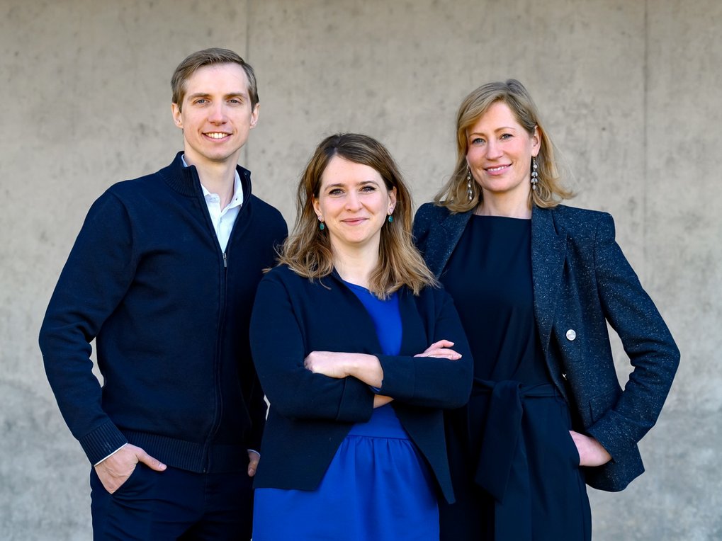 Drei Personen, ein Mann und zwei Frauen, in formeller Kleidung stehen vor einer Betonwand und blicken in die Kamera.