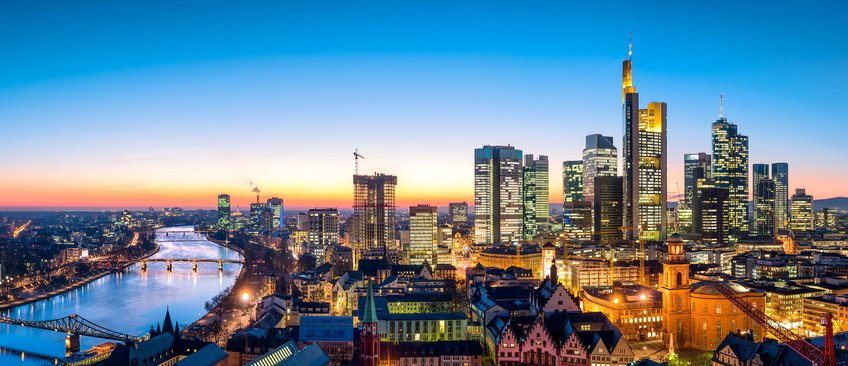 A panoramic view of Frankfurt city centre at sunset, showing numerous illuminated skyscrapers, the River Main and historic buildings in the foreground.